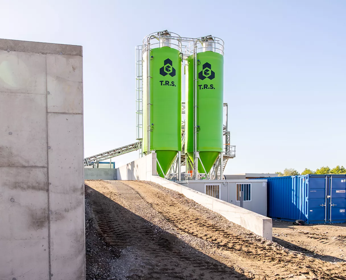 Green silos with recycling symbols beside shipping containers on a dirt road.